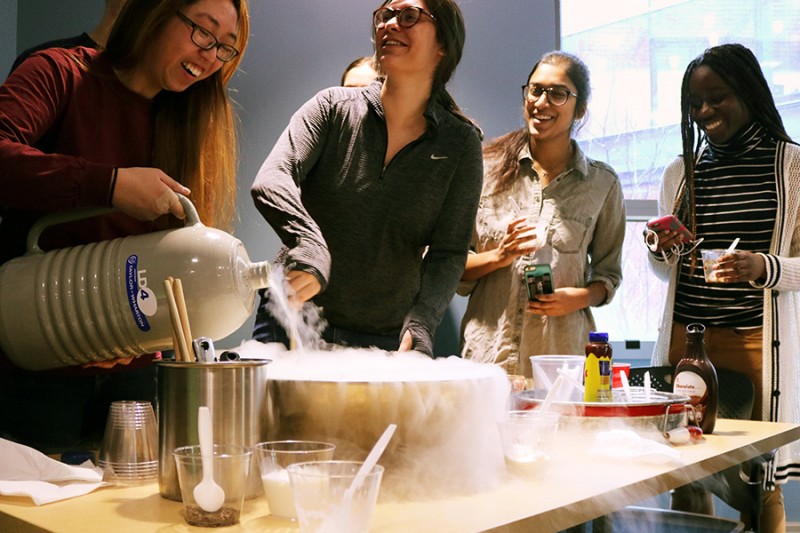 One student pours liquid nitrogen into a bowl of cream while a second stirs. Smoke spills over the bowl that sits on the tan table and two more students watch in the background. 