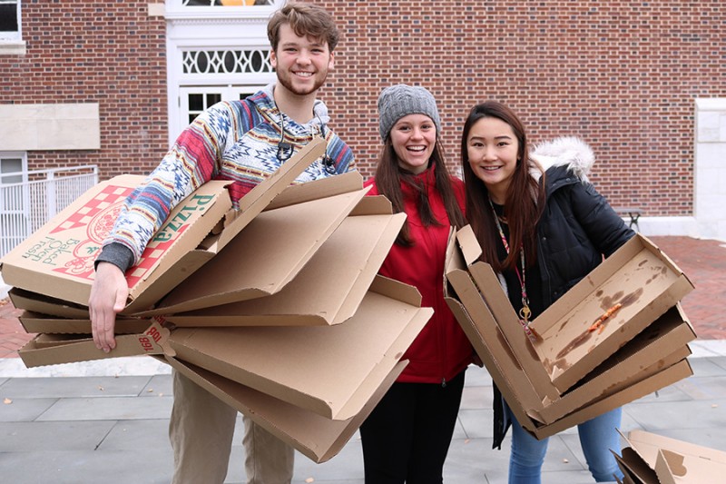 Three students hold empty pizza boxes and smile. They wear winter gear and stand in front of a brick wall.