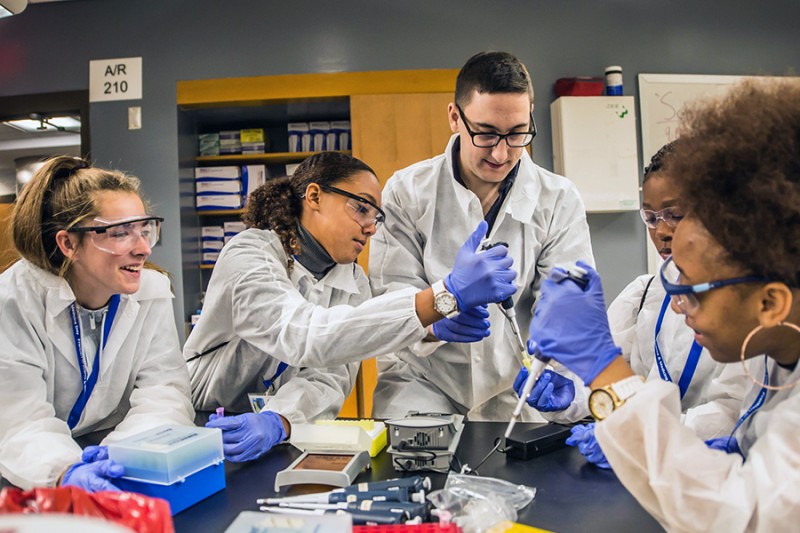 Five students clad in white lab gear and safety glasses wear blue rubber gloves. One girl smiles, they all stand around a table with lab equipment