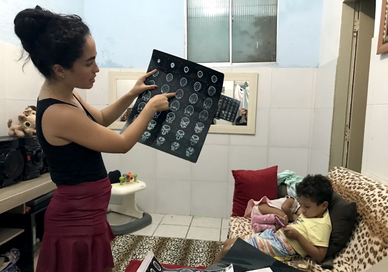 A woman holds a printout of brain scans and points to a spot on the chart while a baby is curled up on a couch next to her and a little boy plays with something in his hand