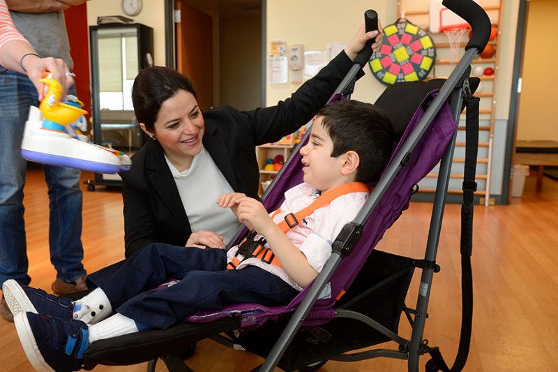 Child sits strapped into a purple stroller and smiles as he is given a toy. A women crouches down next to him and smiles.