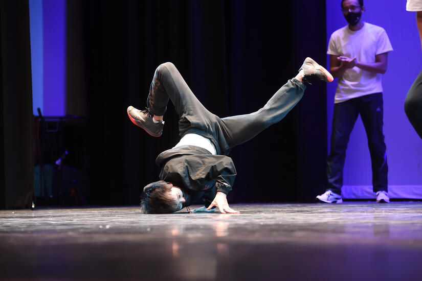 A young adult in a face mask breakdances onstage. Their feet are up in the air.