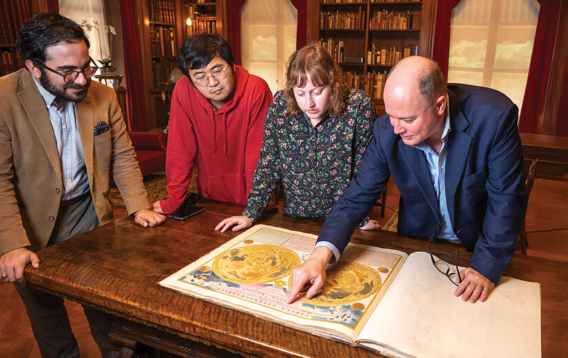 Four people are gathered around a table in an old library, looking at a hand-colored illustration of the moon from 