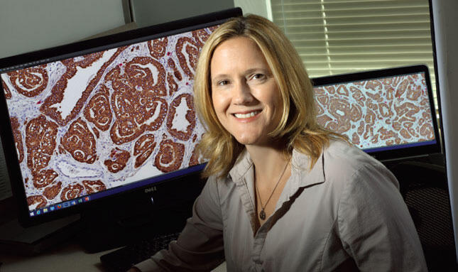 A researcher in a white lab coat sits in front of computer screens showing close-up images of cells