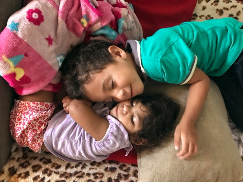 A little girl sleeps while a little boy lays his head down on a pile of pillows and blankets beside her. He smiles.