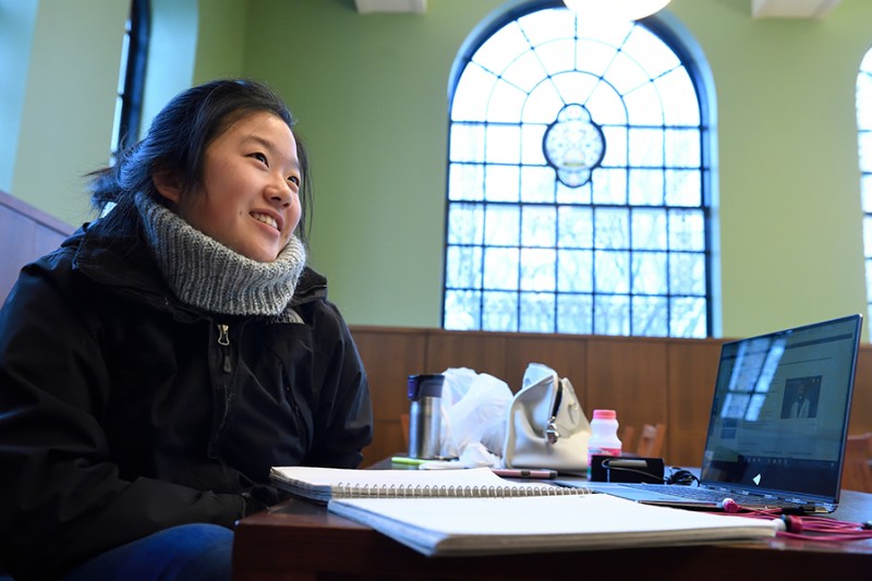 A student wears a scarf and coat and sits a table, a notebook and laptop spread out in front of her