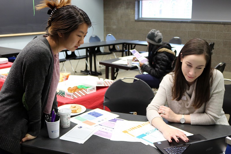 A student leans in to see a computer screen while a staff member show her a website. There are various papers on the table and a box of donuts in the background.