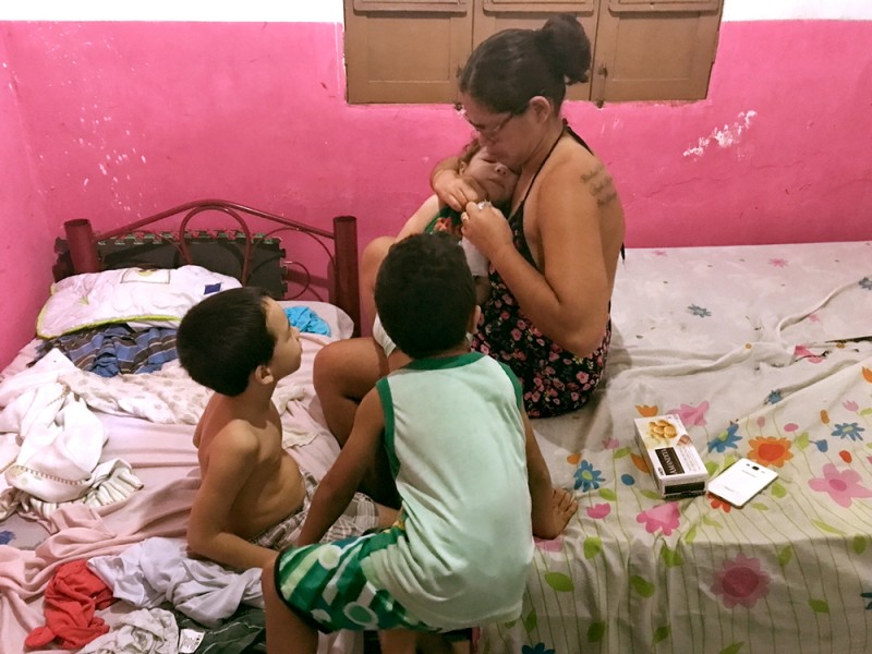 In a room crowded with two beds, a woman holds a baby while she tries to open a package, two little boys watch her