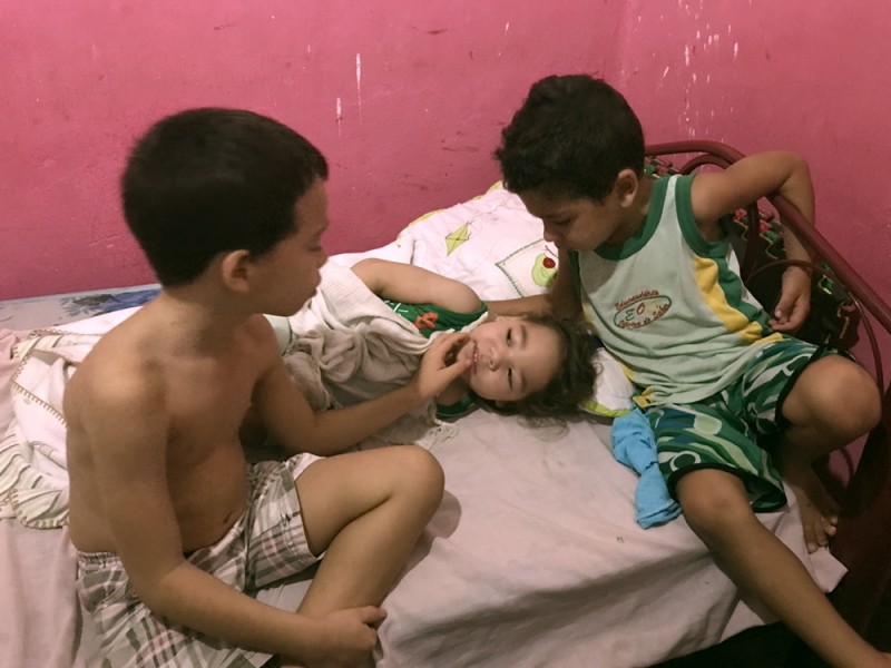 In a room with pink peeling paint, a little girl lays on a bed while two little boys look at her closely