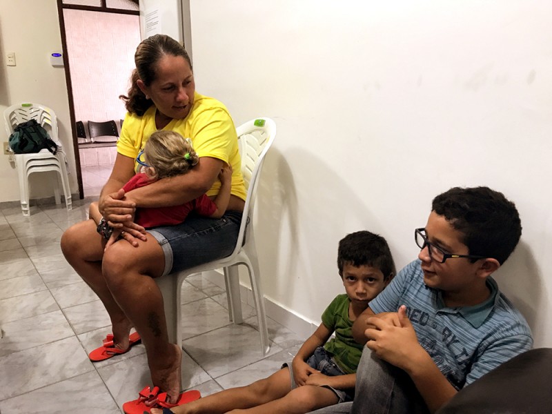A woman holds a baby and sits on a white plastic chair in a kind of waiting room while two little boys sit on the floor; one little boy looks seriously or sadly at the camera
