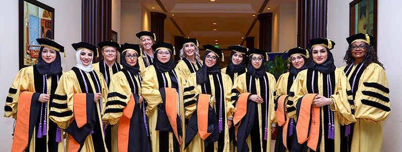 Thirteen women wear commencement regalia, some of whom wear a head covering under their cap
