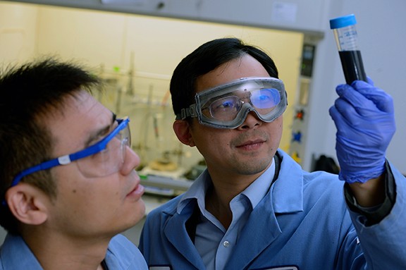 A man wears safety goggles and holds up a test tube with dark liquid and examines it while a second man observes