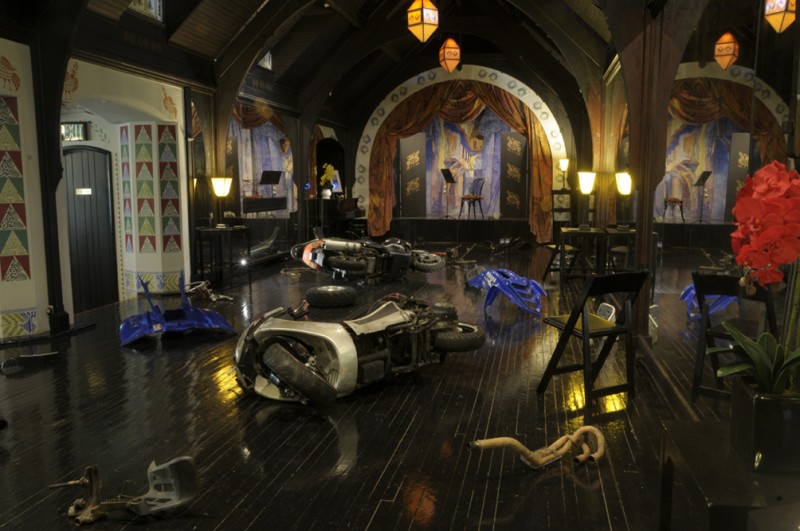 Inside a dimly lit room with gothic features, scooters and dirt bikes are laid on their sides