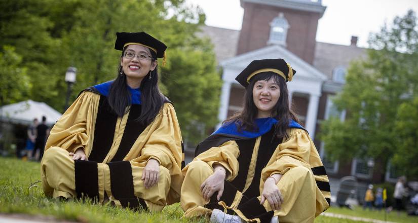 Students in PhD regalia sit in front of Gilman Hall