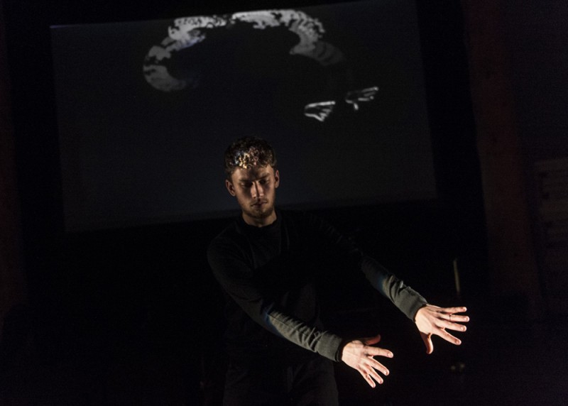 A man in front of a black screen waves his hand in front of a light while smokey swirls are projected on a screen behind him