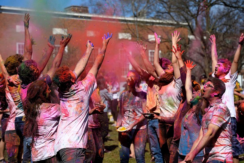 A large group of students wearing clothes covers in colorful powder throw red, green, and pink powder into the air.