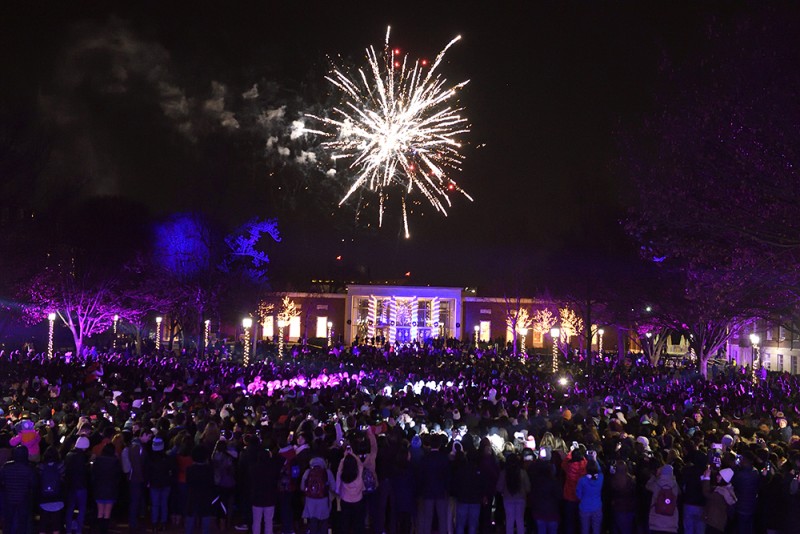 Hundreds of students gather on the quad while the sky lights up with fireworks and laser lights. A campus building in the background glows with holiday lights.