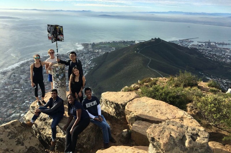 Seven students pose around a flag pole at the top of a mountain while a city and coastline sprawls beneath them
