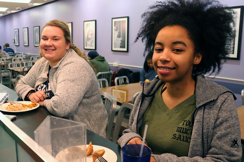 Two students sit next to each other while eating lunch. They both smile in front of a purple wall.