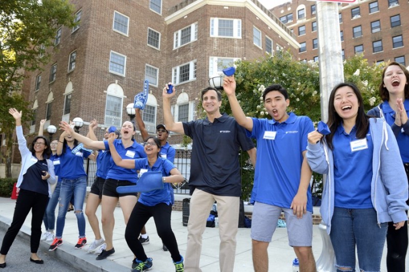 Residence Life staff clad in blue polos excitedly cheer and shake bells as students arrive to campus. JHU President Ronald J. Daniels stands amid them with an equally excited expression.