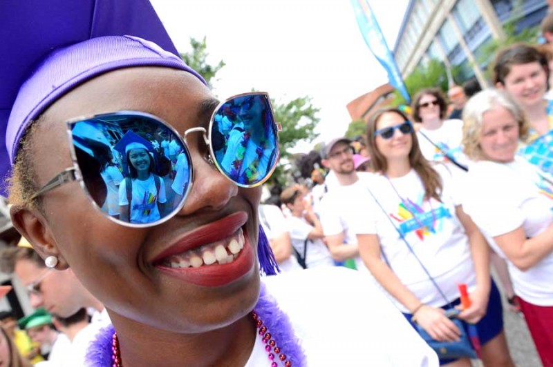 Women wearing purple hat grins with large sunglasses on, in reflection is another woman with a purple graduation hat on and smiling. People in white t-shirt are the background smiling/smirking