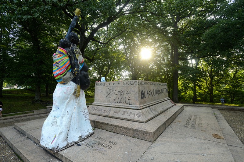 A statue of a woman raising a golden fist in the air and carrying a baby in a sling on her back stands before an empty, vandalized statue base