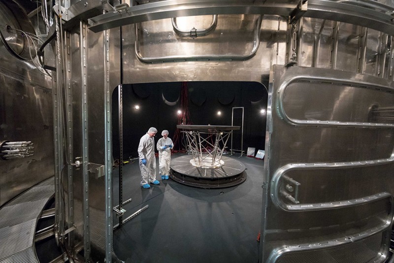 Scientists in lab suits and hair nets stand inside a giant metal vault with a shield bolted to metal brackets inside