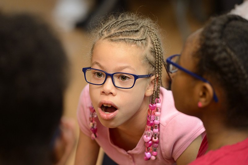 A young girl wearing glasses looks in awe at something outside the line of sight. Her mouth is open in amazement and eyebrows raised.