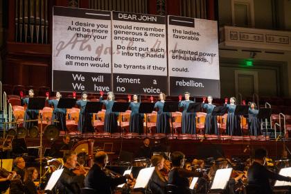 Women stand on stage in front of a giant sign that reads 'Dear John, We will foment a rebellion. We have no voice.'