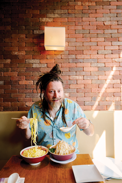 Crazy Legs Conti sits in front of two bowls containing noodles, broth, and sprouts