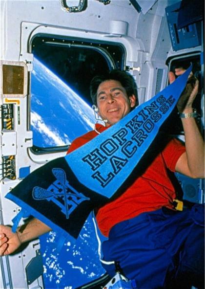 A person in a space capsule, holding a Hopkins Lacrosse flag