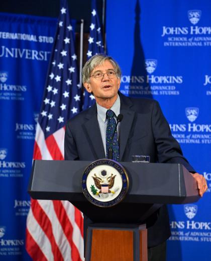 SAIS Dean James Steinberg stands at a podium in front of a blue backdrop; part of an American flag is visible over his right shoulder