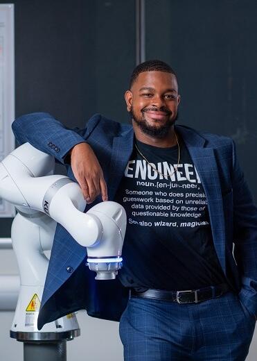 A. Michael West Jr. leans against the white arm of a machine. He is wearing a shirt that reads 