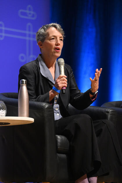 Sarah L. Szanton speaks into a microphone while sitting in a black chair. Next to her is a table with a water bottle.