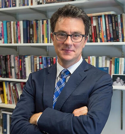 A man wearing glasses stands with his arms folded in front of a large, full bookshelf