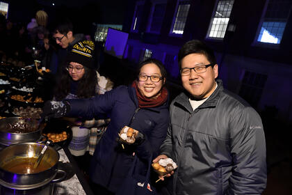 Two adults smile for the camera while standing at a busy outdoor buffet at night.