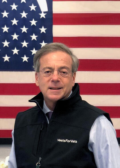 A man wearing a black vest and eyeglasses stands in front of a U.S. flag