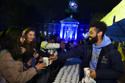 An adult hands out mugs at a booth table. Two college students accept a mug.