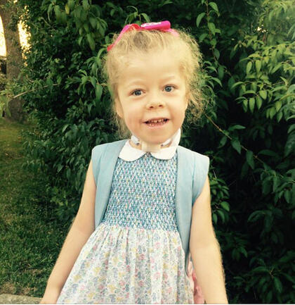 A young girl smiles; she wears a pink bow in her hair, a blue and white dress with floral print, and a backpack