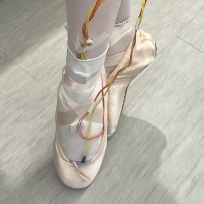 Photo of someone wearing ballet shoes and the PointeSense system, which consists of red and yellow wires running from the wearer's toes to their ankles