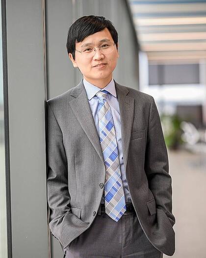 Tinglong Dai leans against a pillar while wearing a grey suit.