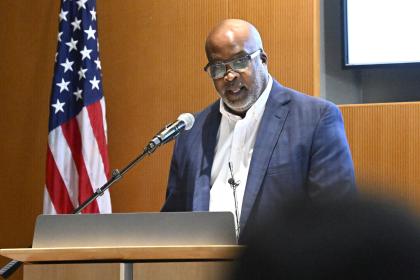 Melvin Wilson speaks into a microphone at a podium. Behind him is an American flag on a flagpole.