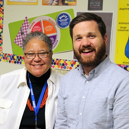 Two people smile in front of a bulletin board plastered with health and wellness posters