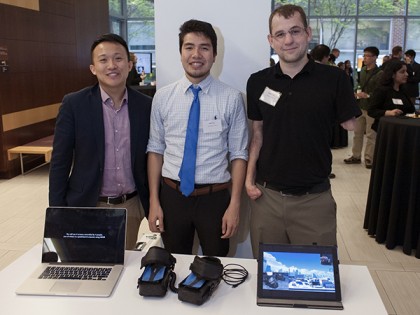 three students stand at a table displaying their design and two computers