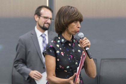 A woman holds a microphone and speaks while a man smiles behind her