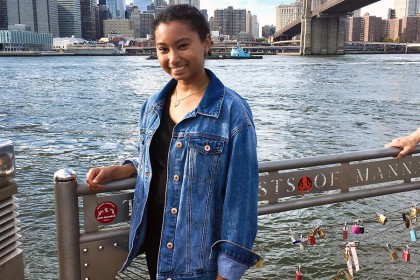 Girl wears jean jacker and smiles while her hand rests on a railing. The city and water is behind her. There are many padlocks on the fencing she leans against. 