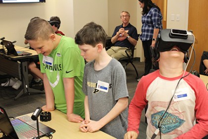 Two boys examine a computer screen while a third boy with VR goggles on his head looks up at the ceiling