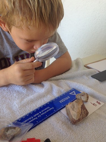 A little boy holds a plastic magnifying glass and inspects a rock that is laid out on a note card labeled with the number 4
