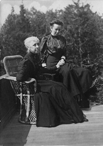 Grainy photograph shows two women, one on the left is sitting while the other stands beside her