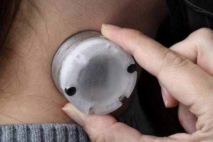 A woman holds a small round device the size of a large bottle cap to her neck